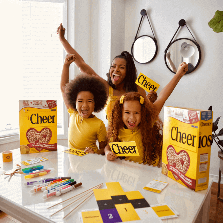 Mom and two kids sitting at a table with their arms up cheering with a Cheerios pack shot