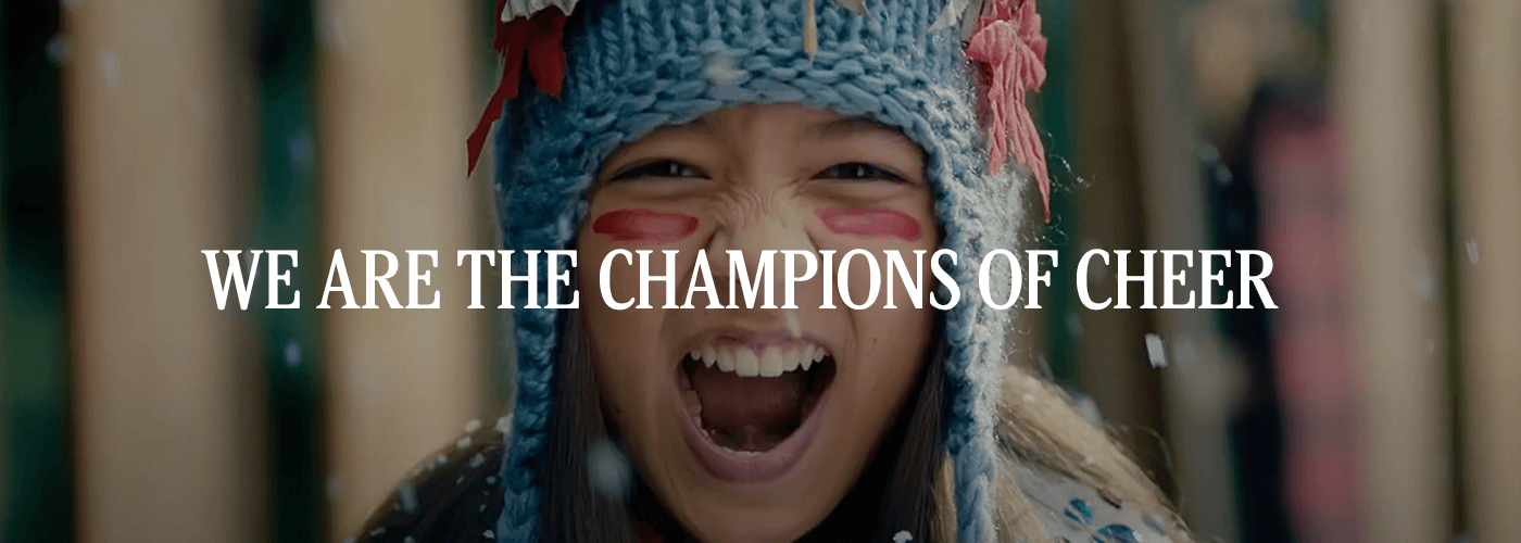 Little girl with blue hat and red warrior paint under her eyes with text across her face saying, "We are the champions of cheer"